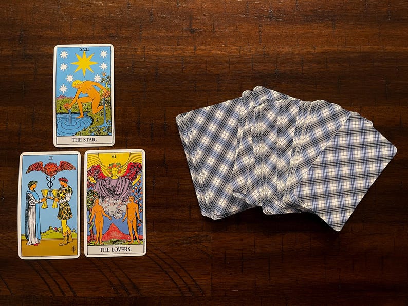 Puede incluir: Una tirada de cartas del tarot con tres cartas boca arriba y una baraja de cartas. Las cartas muestran ilustraciones coloridas con t&iacute;tulos como "The Star" y "The Lovers". La baraja tiene un patr&oacute;n de cuadros azules y blancos en el reverso.