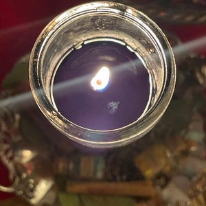 Puede incluir: Una vela morada oscura encendida en un tarro de vidrio transparente. La llama de la vela es brillante contra la cera oscura. El tarro se encuentra sobre una bandeja plateada decorativa, con un fondo borroso de rojo y otros objetos.