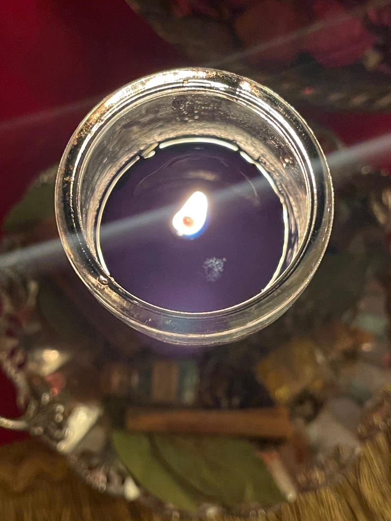 Puede incluir: Una vela morada oscura encendida en un tarro de vidrio transparente. La llama de la vela es brillante contra la cera oscura. El tarro se encuentra sobre una bandeja plateada decorativa, con un fondo borroso de rojo y otros objetos.