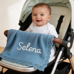 May include: A baby blanket in a stroller. The blue blanket has a waffle weave pattern and a scalloped edge. The name "Selena" is embroidered in white script across the blanket. The baby is wearing a white onesie.