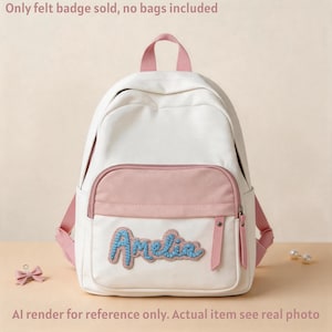 Op de afbeelding: Witte en roze rugzak met de naam "Amelia" in blauwe geborduurde letters. De rugzak heeft roze bandjes en een roze voorvak. De tekst "Only felt badge sold, no bags included" staat bovenaan. De tekst "Al render for reference only. Actual item see real photo" staat onderaan.