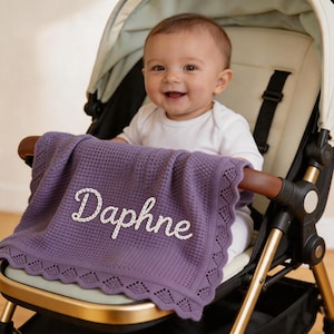 May include: A purple baby blanket with the name "Daphne" embroidered in white script. The blanket has a waffle weave texture and a scalloped edge. The baby is sitting in a stroller.