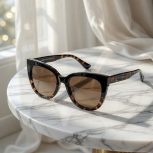 May include: Black cat-eye sunglasses with brown gradient lenses and tortoise-shell arms. The sunglasses are resting on a white marble table. The frame has a thick, bold design.