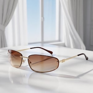 May include: Rimless sunglasses with a gradient brown tint. The frames are gold-colored with brown temples. The sunglasses are resting on a white surface, with a window and curtains in the background.