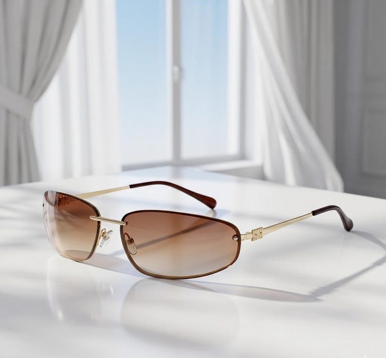 May include: Rimless sunglasses with a gradient brown tint. The frames are gold-colored with brown temples. The sunglasses are resting on a white surface, with a window and curtains in the background.