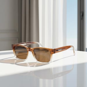 May include: Brown rectangular sunglasses with a transparent frame and tinted lenses. The sunglasses are resting on a white surface, with a window and curtains in the background. The sunglasses have a modern design.