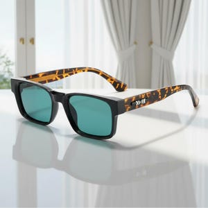 May include: Black-framed sunglasses with green lenses. The temples feature a tortoiseshell pattern. The sunglasses are set on a white surface, with a window and curtains in the background.