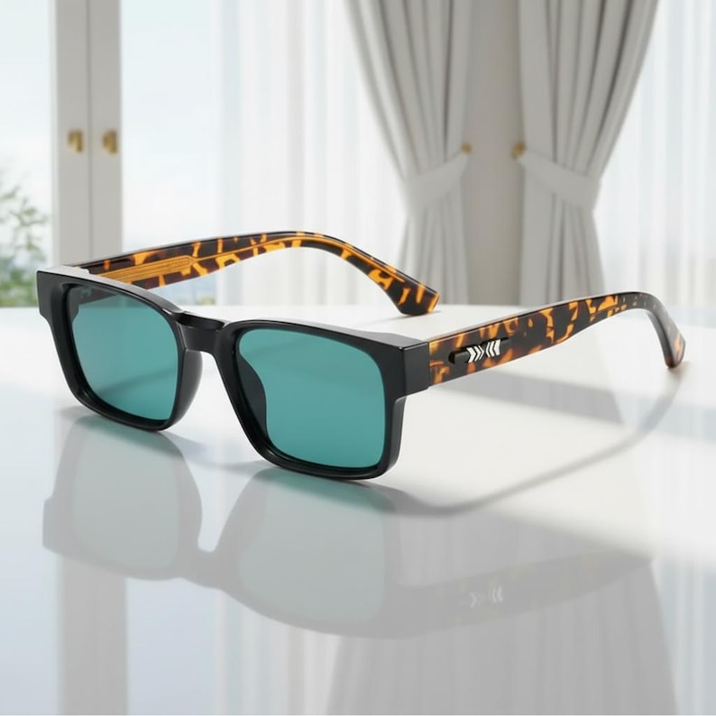 May include: Black-framed sunglasses with green lenses. The temples feature a tortoiseshell pattern. The sunglasses are set on a white surface, with a window and curtains in the background.