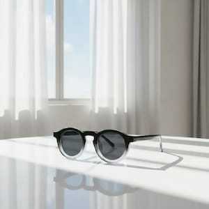 Könnte beinhalten: Runde Sonnenbrille mit schwarzem Rahmen und dunklen Gläsern liegt auf einer weißen Oberfläche. Der untere Teil des Rahmens ist durchscheinend. Im Hintergrund ein Fenster mit weißen Vorhängen und blauem Himmel.
