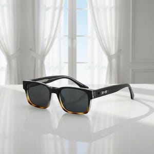 May include: Black sunglasses with a rectangular frame and gradient brown-tinted lower edges. The temples are black with a silver arrow design. The lenses are dark gray, and the sunglasses are set on a white surface.