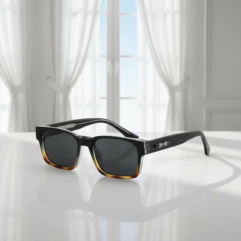 May include: Black sunglasses with a rectangular frame and gradient brown-tinted lower edges. The temples are black with a silver arrow design. The lenses are dark gray, and the sunglasses are set on a white surface.