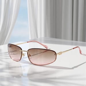 May include: Rimless sunglasses with a gradient brown and pink tint. The frames are gold with pink temples. The sunglasses are on a white surface with a soft background.
