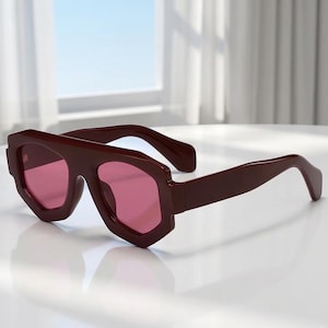 Puede incluir: Gafas de sol color burdeos con montura geométrica y lentes rosas. Las gafas tienen un diseño grueso y angular, con un puente y patillas que se extienden hacia los lados. Las lentes son de un color rosa vibrante, ofreciendo un contraste con la montura oscura.