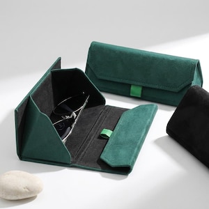 Peut inclure: Un ensemble d'étuis à lunettes triangulaires pliables en matière façon daim vert foncé. Un étui est ouvert, révélant un intérieur noir et une paire de lunettes à monture sombre. Un deuxième étui vert fermé et un étui noir partiel sont visibles.