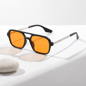 Peut inclure: Lunettes de soleil de style aviateur avec des verres teintés orange vif dans une monture noire épaisse. Les branches sont en métal argenté fin, se terminant par des embouts en plastique noir. Le pont présente un design à double barre.
