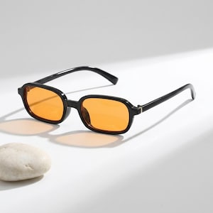 Peut inclure: Lunettes de soleil dotées d'une monture en plastique noir brillant et de verres rectangulaires. Les verres sont d'une couleur orange translucide et vive. Les branches sont fines et noires, reliées à la monture par de petites charnières métalliques.