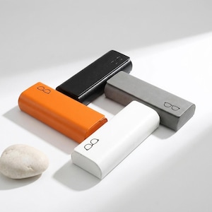 May include: Four rectangular glasses cases in black, orange, grey, and white, each featuring a small black line-art icon of spectacles printed on the top surface. The cases are arranged in a cluster alongside a single smooth, light-coloured stone.