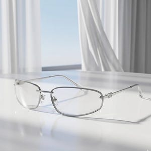 May include: Silver-framed eyeglasses with a rectangular lens shape. The glasses have a thin, minimalist design with a bridge and temples. The lenses are clear, and the frame is reflective. The glasses are resting on a white surface.