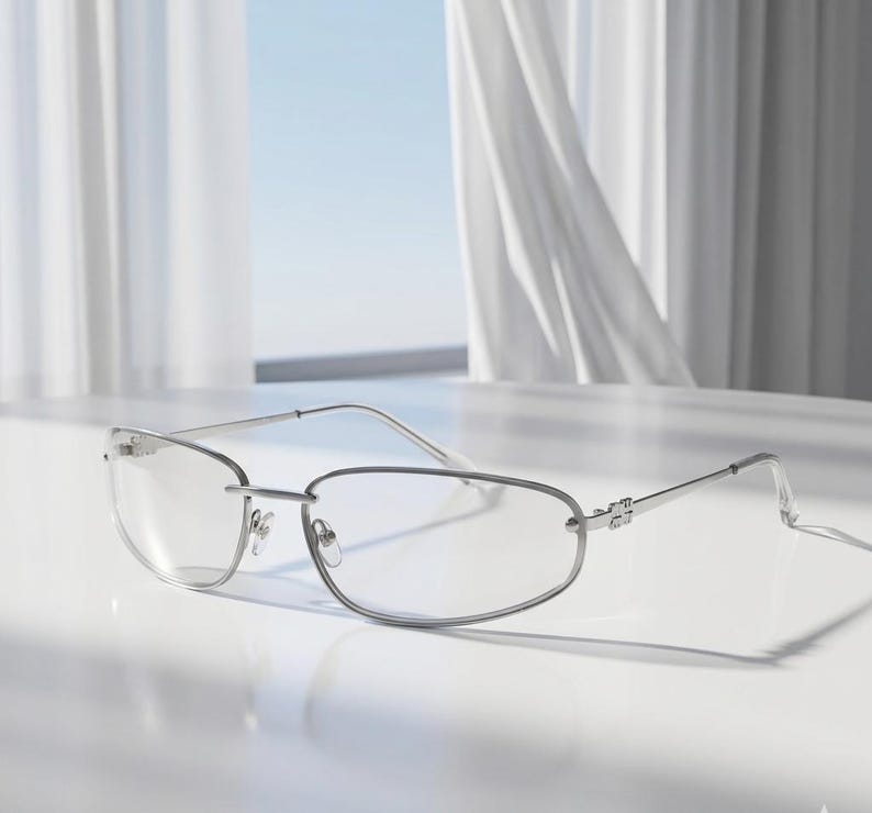 May include: Silver-framed eyeglasses with a rectangular lens shape. The glasses have a thin, minimalist design with a bridge and temples. The lenses are clear, and the frame is reflective. The glasses are resting on a white surface.
