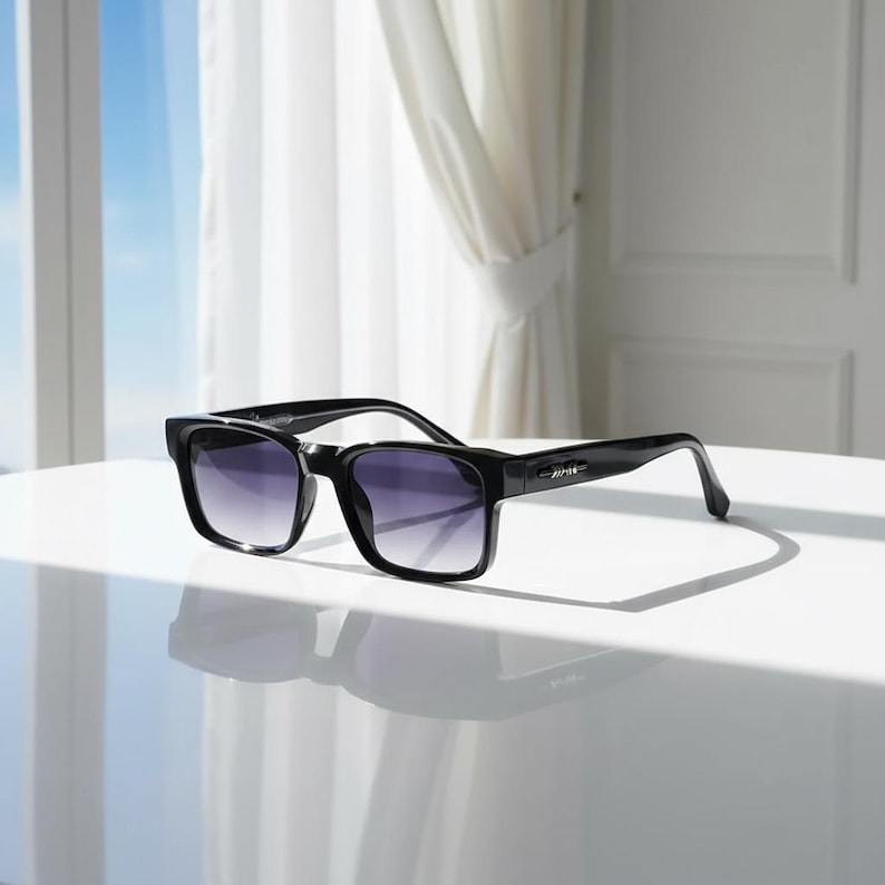 May include: Black square-framed sunglasses with gradient lenses. The sunglasses are on a white surface, with a window and curtains in the background. The lenses transition from dark at the top to lighter at the bottom.