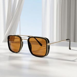 Puede incluir: Gafas de sol cuadradas con montura negra y dorada y lentes ámbar. Las gafas tienen un doble puente y un diseño moderno. Las lentes son de color ámbar y la montura es negra y dorada.