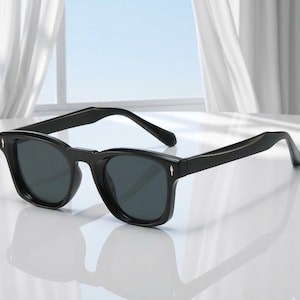 May include: Black sunglasses with dark lenses. The sunglasses have a thick, rectangular frame with a subtle arrow detail on the temples. The glasses are set against a bright, neutral background.
