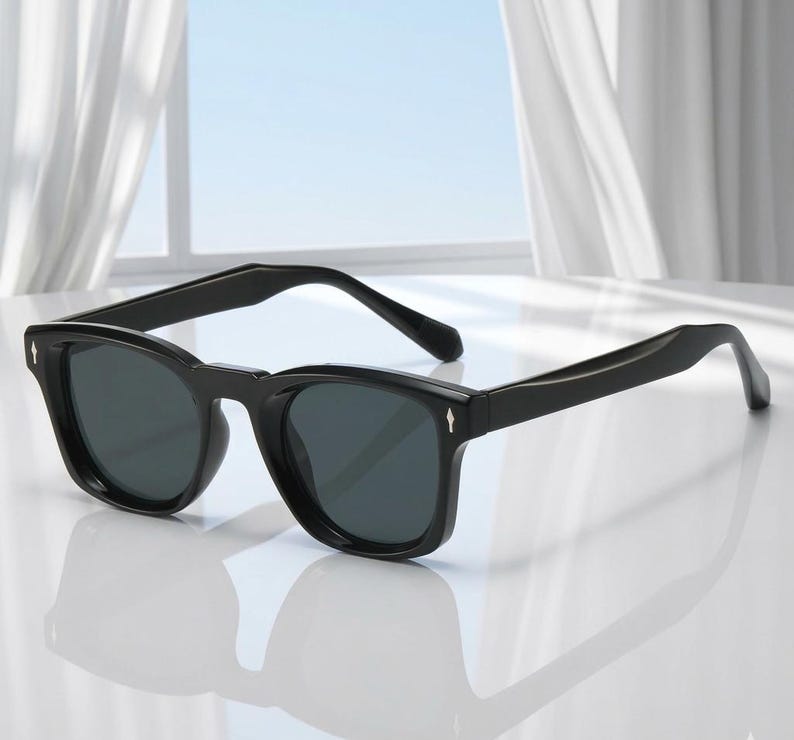 May include: Black sunglasses with dark lenses. The sunglasses have a thick, rectangular frame with a subtle arrow detail on the temples. The glasses are set against a bright, neutral background.