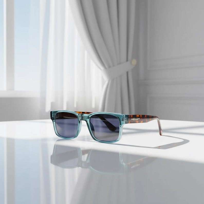 May include: Square-framed sunglasses with light blue frames and dark lenses. The temples have a tortoiseshell pattern. The sunglasses are resting on a white surface, with a window and curtains in the background.