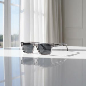 May include: Clear rectangular sunglasses with dark gray lenses rest on a white surface. The frames are transparent, and the sunglasses cast a shadow. The design is modern and minimalist, suitable for eye protection.