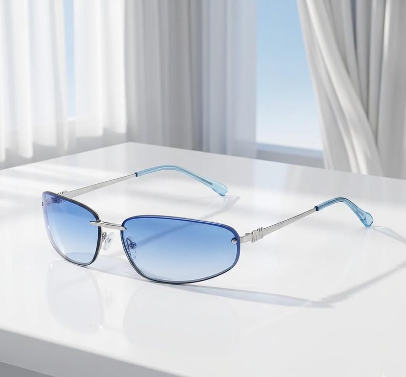 May include: Rimless sunglasses with a gradient blue tint. The frames are silver with light blue temples. The lenses are a rectangular shape. The sunglasses are sitting on a white surface.