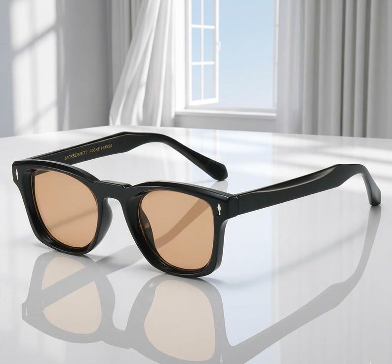 May include: Black sunglasses with square frames and light brown lenses. The sunglasses have a classic design with a modern twist. The text "JACKIE WITT FORME DESIGN" is printed on the top of the frame.