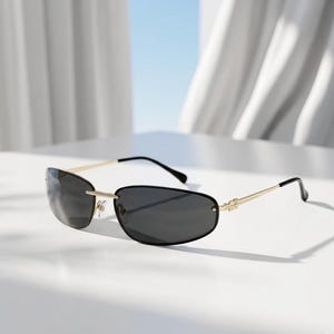 May include: Rimless sunglasses with dark lenses and gold-colored metal frames. The sunglasses have a sleek, modern design with a thin frame and curved temples. The lenses are a dark gray color, providing shade and style.