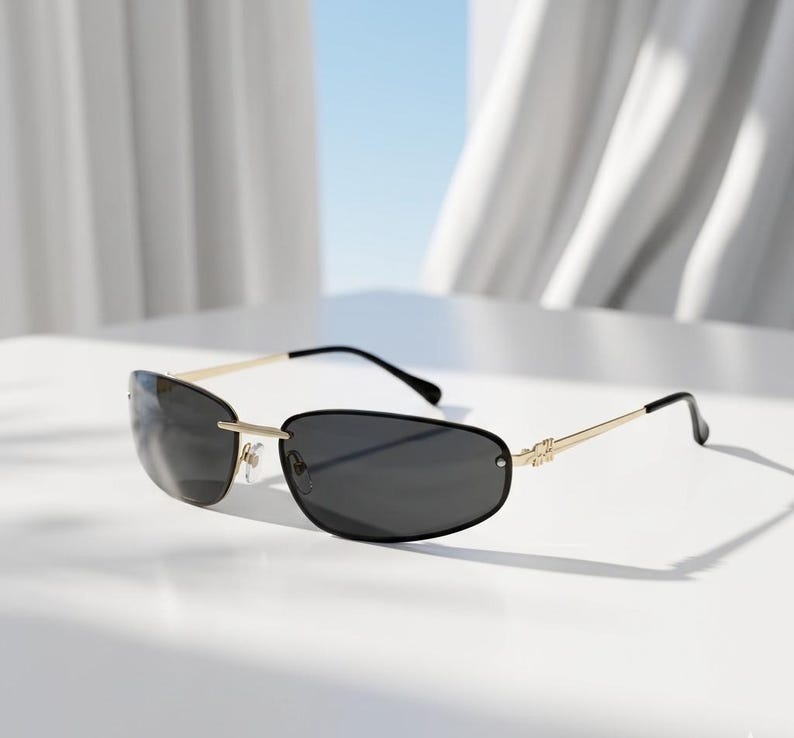 May include: Rimless sunglasses with dark lenses and gold-colored metal frames. The sunglasses have a sleek, modern design with a thin frame and curved temples. The lenses are a dark gray color, providing shade and style.