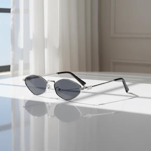 May include: Silver-framed sunglasses with a distinctive geometric design and dark grey lenses. The sunglasses feature black temples and are set against a white backdrop, offering a modern and stylish appearance. These sunglasses are a fashion accessory.