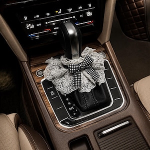 May include: A decorative gear shift cover featuring a white floral lace ruffle and a black and white gingham bow. The accessory is positioned around the base of a black and silver gear shifter in a vehicle interior with wood grain trim and buttons.