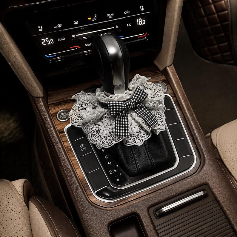 May include: A decorative gear shift cover featuring a white floral lace ruffle and a black and white gingham bow. The accessory is positioned around the base of a black and silver gear shifter in a vehicle interior with wood grain trim and buttons.