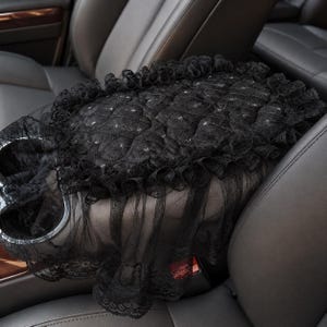May include: A black car center console cover featuring a quilted texture with small embedded rhinestones. The rectangular cover is edged with a ruffled lace trim and a sheer black tulle overlay that drapes down the sides of the console.