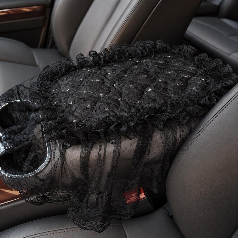 May include: A black car center console cover featuring a quilted texture with small embedded rhinestones. The rectangular cover is edged with a ruffled lace trim and a sheer black tulle overlay that drapes down the sides of the console.