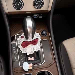 May include: A car gear shift lever adorned with a white lace collar featuring a deep red bow and a central pearl accent. Below the collar, the black leather shift boot is decorated with a small pink bow and a light blue cloud-shaped charm.