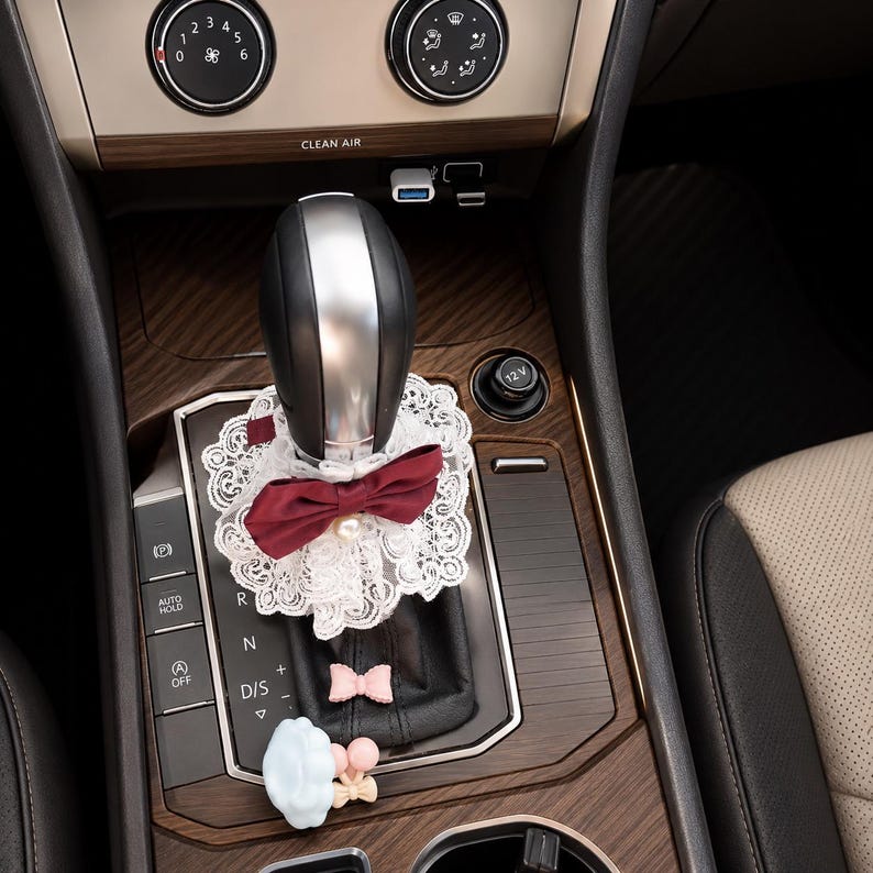 May include: A car gear shift lever adorned with a white lace collar featuring a deep red bow and a central pearl accent. Below the collar, the black leather shift boot is decorated with a small pink bow and a light blue cloud-shaped charm.