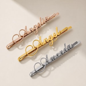 May include: Three personalised hair slides in rose gold, gold, and silver. Each slide features a name in a cursive font: Angelia, Angel, and Amanda. The slides are displayed on a light-coloured surface.