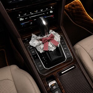 May include: A decorative white lace collar with a red and white gingham bow wrapped around the black leather gear shift knob of a vehicle. The gear shift is positioned on a dark wood grain center console with various control buttons and digital displays.
