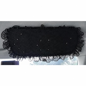 May include: A black car sun visor cover featuring a delicate lace overlay with ruffled edges and small, scattered rhinestone embellishments. The rectangular fabric accessory is designed to attach to a vehicle's interior sun visor mechanism.