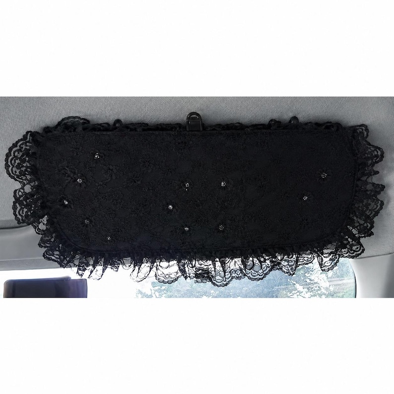 May include: A black car sun visor cover featuring a delicate lace overlay with ruffled edges and small, scattered rhinestone embellishments. The rectangular fabric accessory is designed to attach to a vehicle's interior sun visor mechanism.