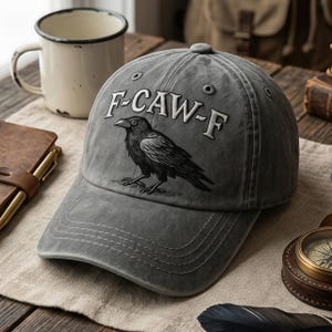 May include: Gray baseball cap with the text "F-CAW-F" above a black crow illustration. The cap has a curved brim and a button on top. The hat is made of a soft, worn material.