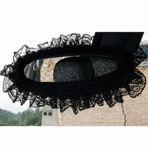 May include: A black lace cover for a vehicle rearview mirror, featuring a ruffled edge design that frames the mirror. The delicate, semi-sheer fabric has a floral pattern and is secured around the perimeter of the mirror housing.