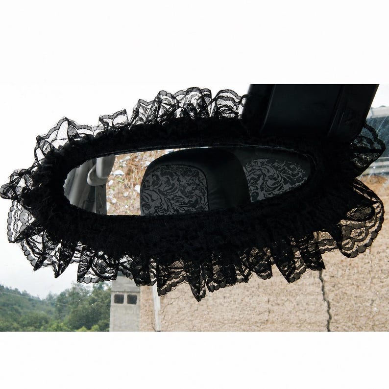 May include: A black lace cover for a vehicle rearview mirror, featuring a ruffled edge design that frames the mirror. The delicate, semi-sheer fabric has a floral pattern and is secured around the perimeter of the mirror housing.
