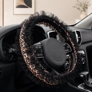 May include: A steering wheel cover featuring a brown and black leopard print pattern, accented with a layered black tulle ruffle trim along the outer edge. The cover is fitted onto a black steering wheel inside a vehicle dashboard environment.