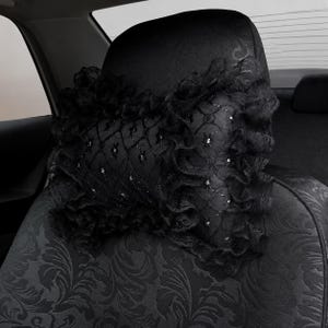 May include: A black car headrest pillow featuring a decorative lace overlay with a diamond grid pattern and ruffled edges. The pillow is secured to a black car seat that has a subtle, dark-toned floral jacquard fabric pattern throughout.