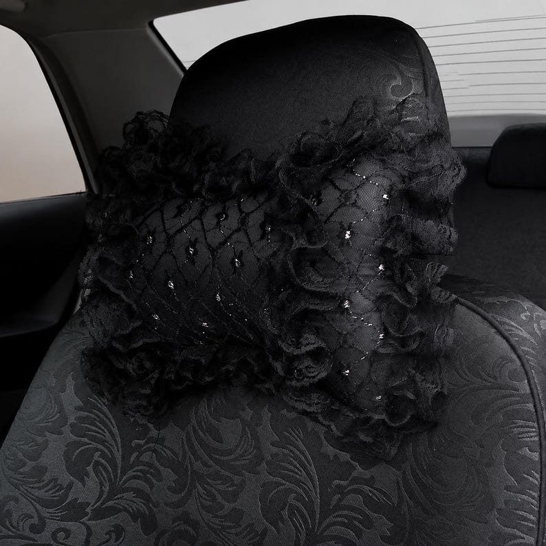 May include: A black car headrest pillow featuring a decorative lace overlay with a diamond grid pattern and ruffled edges. The pillow is secured to a black car seat that has a subtle, dark-toned floral jacquard fabric pattern throughout.